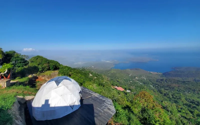 Domo geodésico blanco sobre plataforma elevada en la cima de una montaña, con vista panorámica al mar azul, bahía y selva verde bajo cielo despejado.