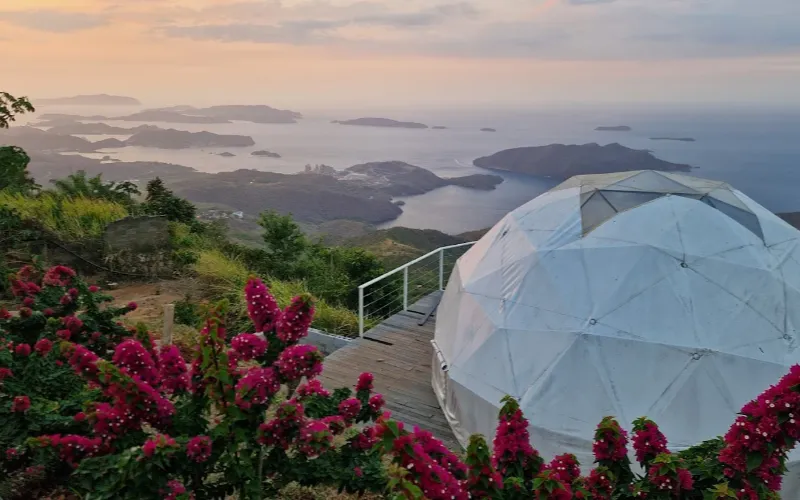 Domo geodésico blanco rodeado de buganvilias fucsia en primer plano, con deck de madera y vista al atardecer sobre archipiélago de islas y mar en tonos rosados.