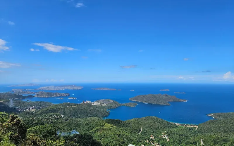 Vista panorámica desde la cima hacia un archipiélago de islas verdes sobre mar azul intenso, con ciudad costera y vegetación tropical bajo cielo azul despejado.