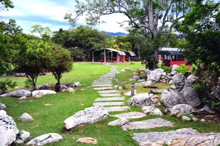 Sendero de piedras irregulares atravesando jardín verde con árboles, rocas y flores rojas, conduciendo hacia estructura roja del glamping con montañas al fondo.