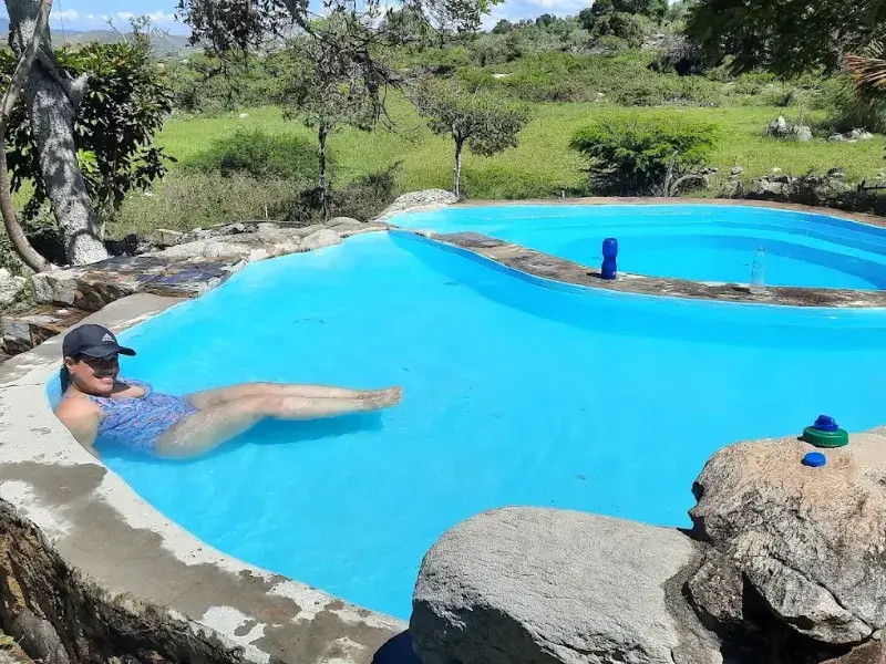 Persona relajándose en piscina de forma orgánica de agua azul turquesa, rodeada de rocas naturales y vegetación silvestre bajo cielo nublado.