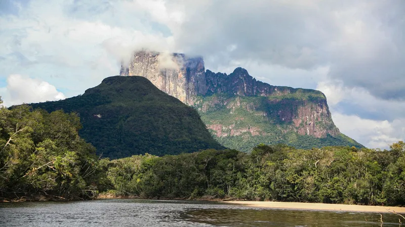 Tepuy de paredes verticales de roca oscura cubierto parcialmente por nubes, rodeado de densa selva tropical amazónica y un río de aguas tranquilas en primer plano, en el Parque Nacional Canaima, Venezuela.