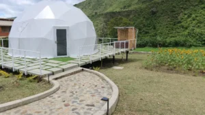 Domo geodésico blanco con entrada principal, deck de madera con barandales blancos y camino de piedra, rodeado de girasoles y vegetación montañosa al fondo.