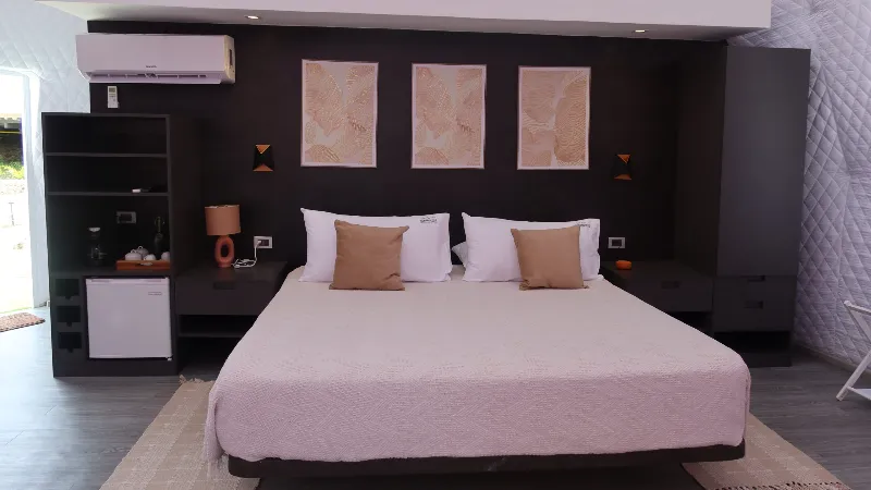 Habitación de glamping con cama doble de ropa blanca, cabecera oscura con tres cuadros decorativos, aire acondicionado, minibar y lámpara de mesa en tono terracota.