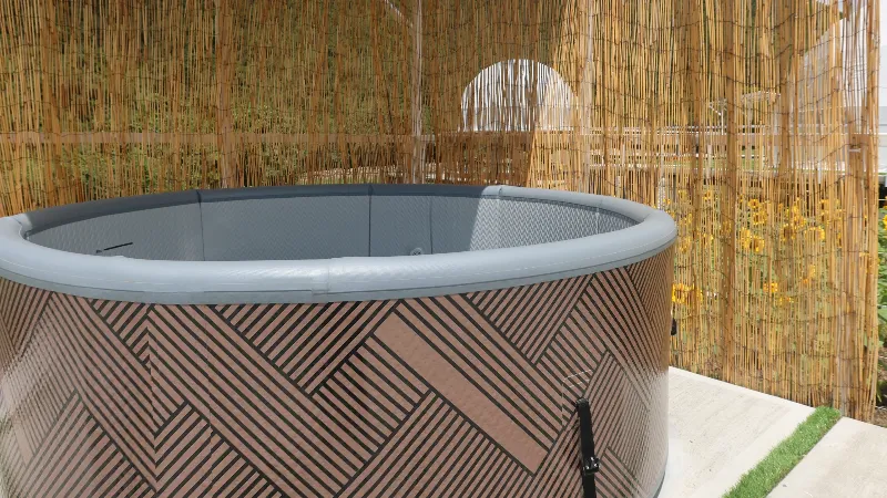 Jacuzzi circular exterior de color gris y madera, ubicado dentro de un espacio privado delimitado por biombos de bambú, con girasoles visibles al fondo.