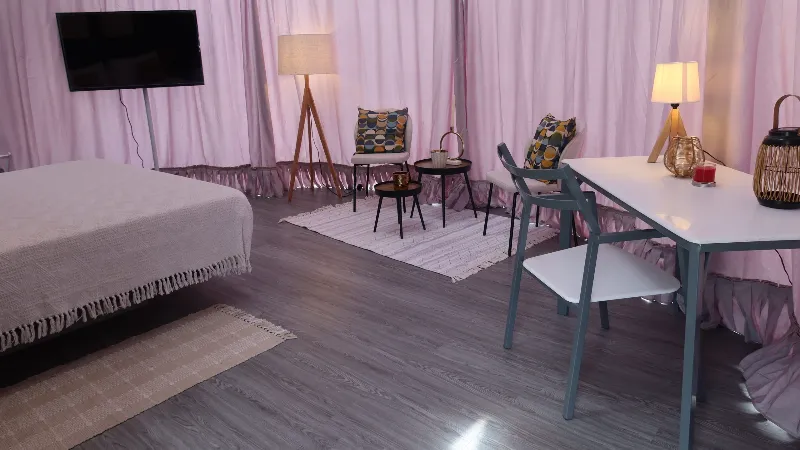Interior del domo con cama, mesa de comedor blanca, sillas, área de descanso con sillones, televisor de pared y cortinas rosas que recubren las paredes curvas.