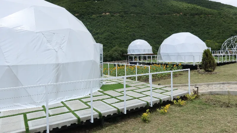 Vista panorámica del glamping Casa Carú con tres domos geodésicos blancos alineados sobre decks con barandales, jardín de girasoles y montañas verdes de fondo.