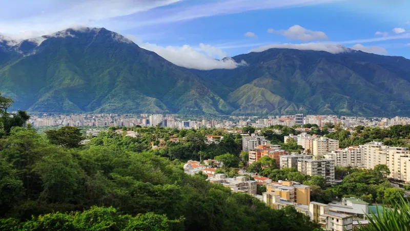 Vista panorámica de Caracas con una extensa área urbana de edificios residenciales y vegetación exuberante, enmarcada por la imponente cadena montañosa del Ávila con sus cumbres entre nubes, Venezuela.