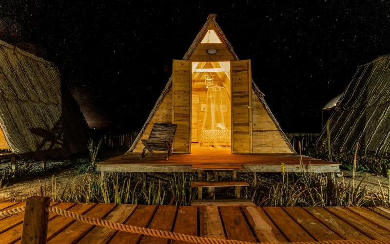 Cabaña A-frame iluminada por dentro durante la noche estrellada en el glamping Cubagua Lodge, isla de Cubagua, Nueva Esparta, Venezuela