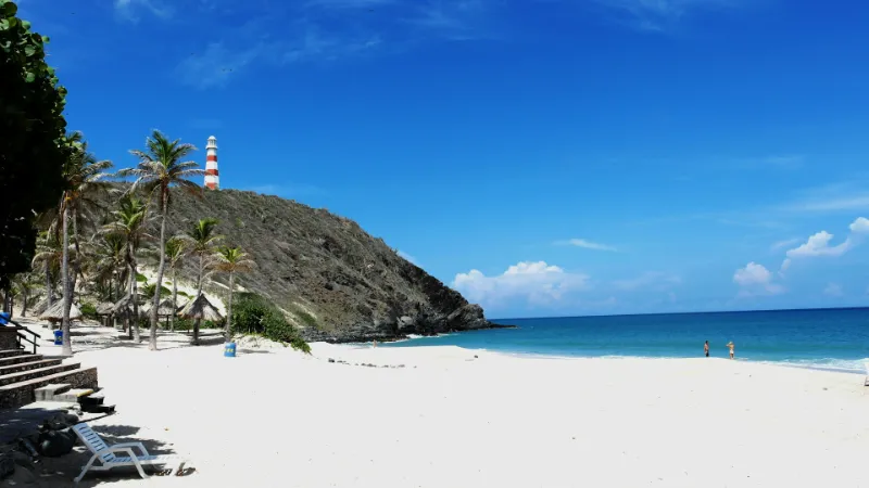 Playa de arena blanca y aguas turquesas con palmeras, un faro de franjas rojas y blancas sobre un cerro rocoso al fondo, y dos personas caminando a orillas del mar bajo un cielo azul despejado, en Nueva Esparta, Venezuela.