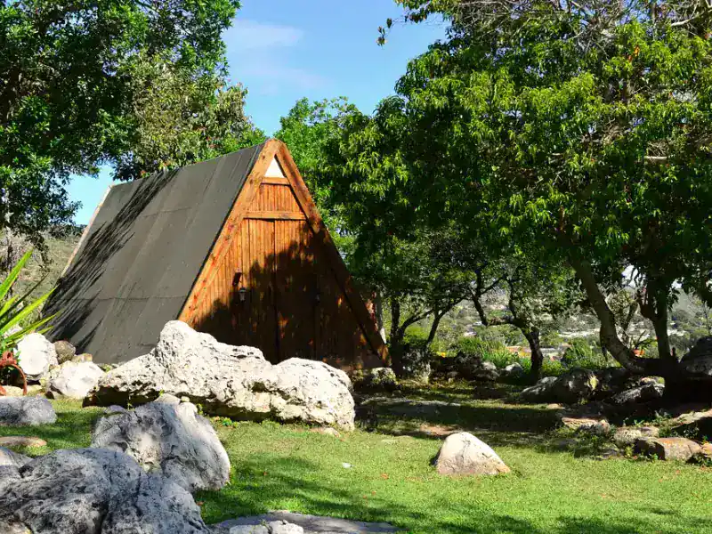 Carpa tipo A-frame con fachada de madera y techo de lona oscura, rodeada de árboles frondosos y rocas en jardín verde bajo cielo azul.