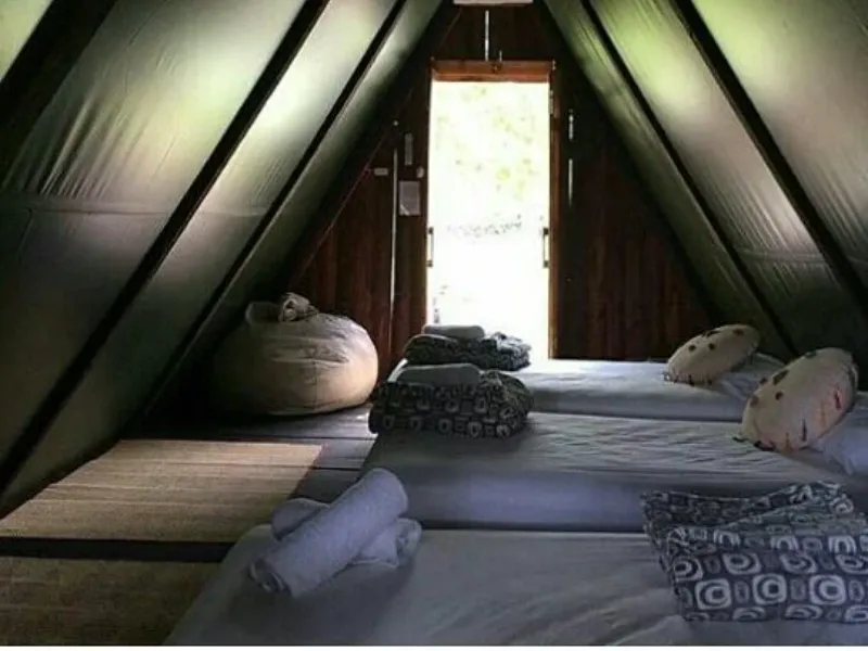 Interior triangular de carpa glamping con tres colchonetas, toallas enrolladas, puf y puerta de madera abierta con luz natural entrando.