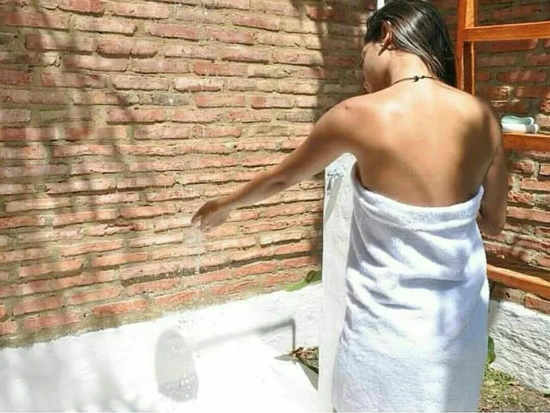 Persona envuelta en toalla blanca probando el agua de una bañera exterior junto a pared de ladrillo y escalera de madera al sol.