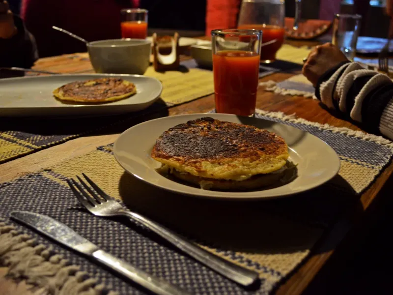 Desayuno con arepa o tortilla en plato blanco, jugo de tomate y cubiertos sobre mantel artesanal en mesa de madera rústica.