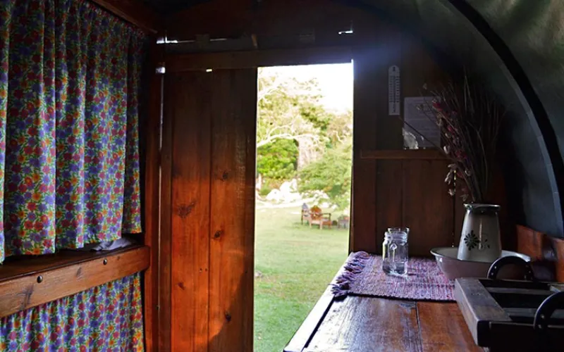 Interior de carreta con mesa de madera, cortinas florales de colores, puerta abierta con vista al jardín iluminado por luz natural.