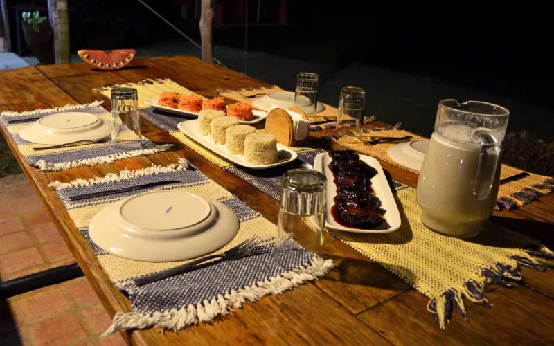 Mesa de madera rústica servida de noche con platos, costillas glaseadas, arroz, frutas, jarra de bebida y mantelería artesanal.