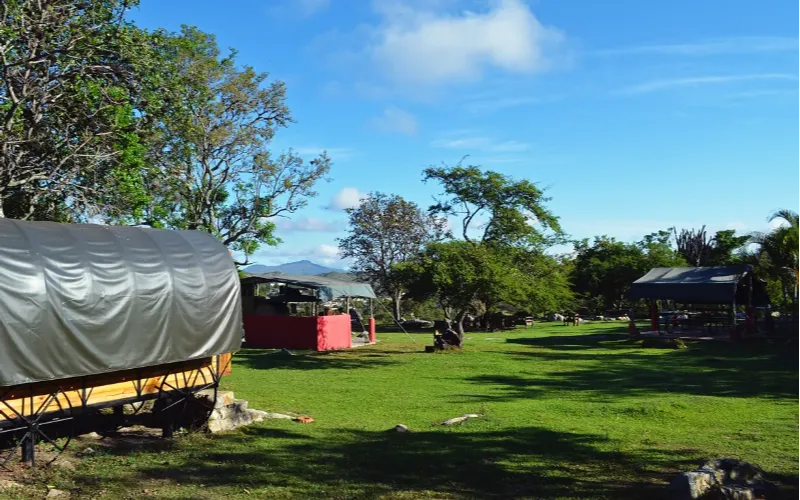 Vista panorámica del glamping con carretas de madera distribuidas en amplia zona verde, árboles frondosos y montañas al fondo bajo cielo azul.