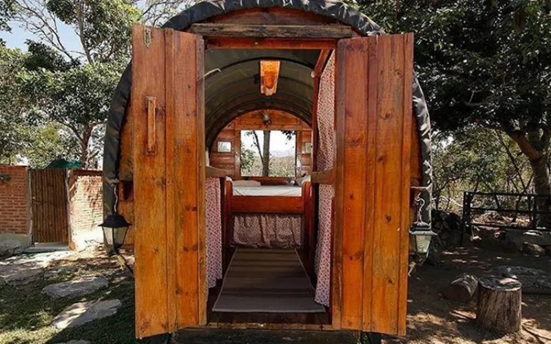 Puerta doble de madera abierta mostrando el interior rústico de una carreta glamping con cama al fondo y techo arqueado.