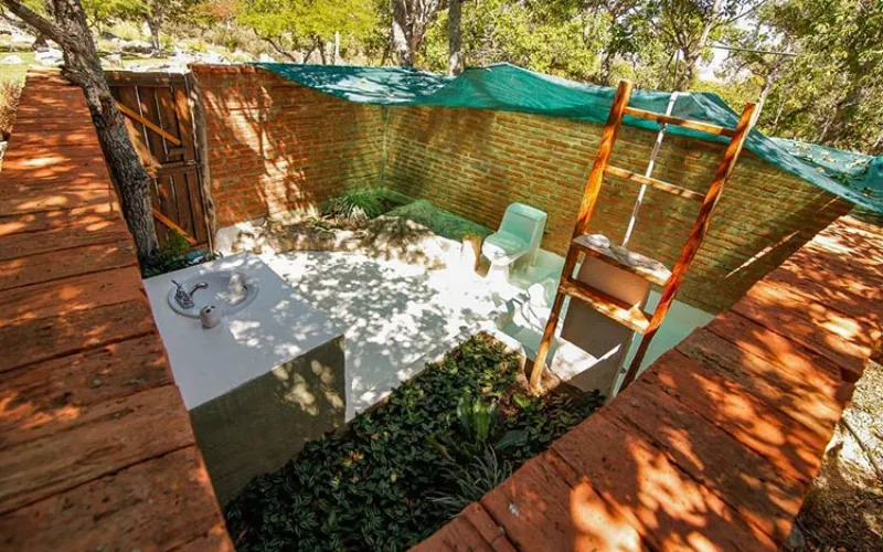 Baño exterior descubierto con paredes de ladrillo, lavamanos blanco, inodoro y escalera de madera bajo malla de sombra verde.