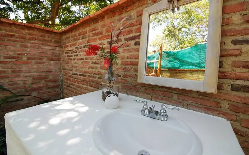 Lavamanos blanco exterior con espejo de madera, florero con flores rojas y paredes de ladrillo a cielo abierto.