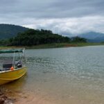 Bote amarillo con toldo atracado a orilla arenosa del embalse, con bosque tropical y montañas al fondo