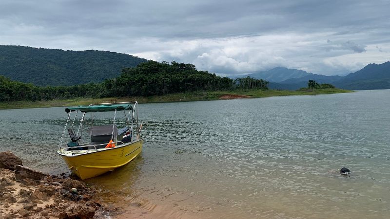 Bote amarillo con toldo atracado a orilla arenosa del embalse, con bosque tropical y montañas al fondo