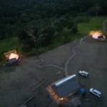 Vista aérea nocturna de dos bell tents iluminadas entre selva y sendero, con cabaña de paja y vehículos estacionados