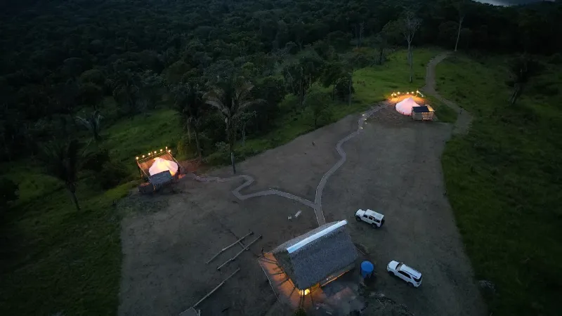 Vista aérea nocturna de dos bell tents iluminadas entre selva y sendero, con cabaña de paja y vehículos estacionados
