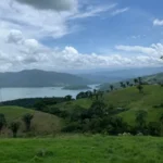 Paisaje de colinas verdes onduladas con embalse al fondo, torres eléctricas y cielo con nubes blancas