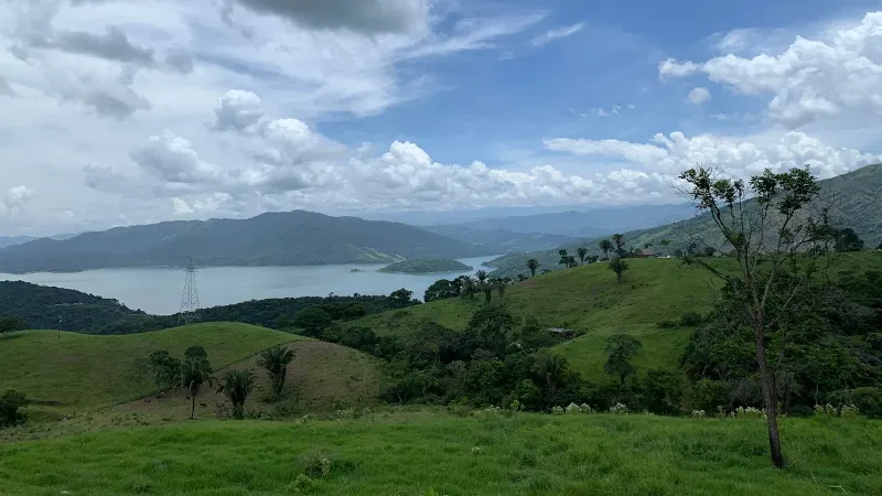 Paisaje de colinas verdes onduladas con embalse al fondo, torres eléctricas y cielo con nubes blancas