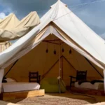 Exterior de bell tent beige abierta mostrando dos camas individuales con sillas de madera tallada y lámparas colgantes