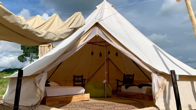 Exterior de bell tent beige abierta mostrando dos camas individuales con sillas de madera tallada y lámparas colgantes