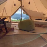 Interior de bell tent con mecedora, puf verde, camas a los lados y vista al embalse y montañas desde la entrada abierta
