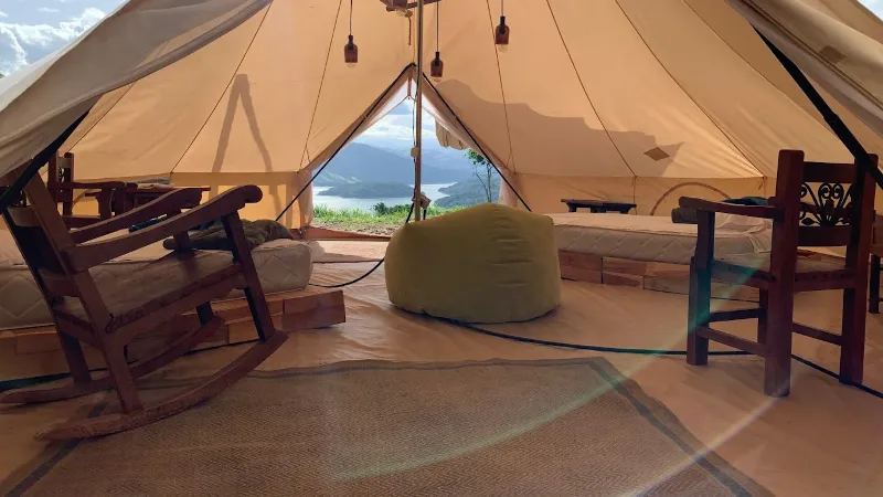 Interior de bell tent con mecedora, puf verde, camas a los lados y vista al embalse y montañas desde la entrada abierta