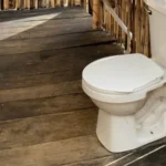 Baño rústico con sanitario blanco sobre piso de madera, rodeado de paredes de guadua en corredor al aire libre