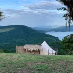 Bell tent blanca y cabaña de techo de paja en colina verde, flanqueadas por palmas, con embalse y montañas al fondo