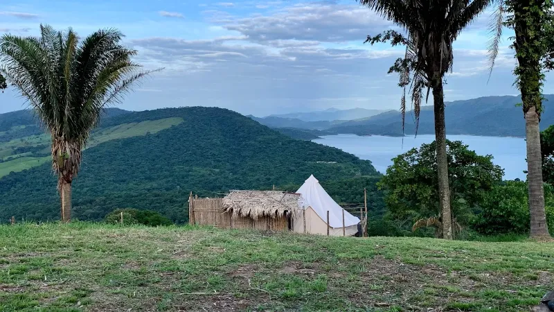 Bell tent blanca y cabaña de techo de paja en colina verde, flanqueadas por palmas, con embalse y montañas al fondo