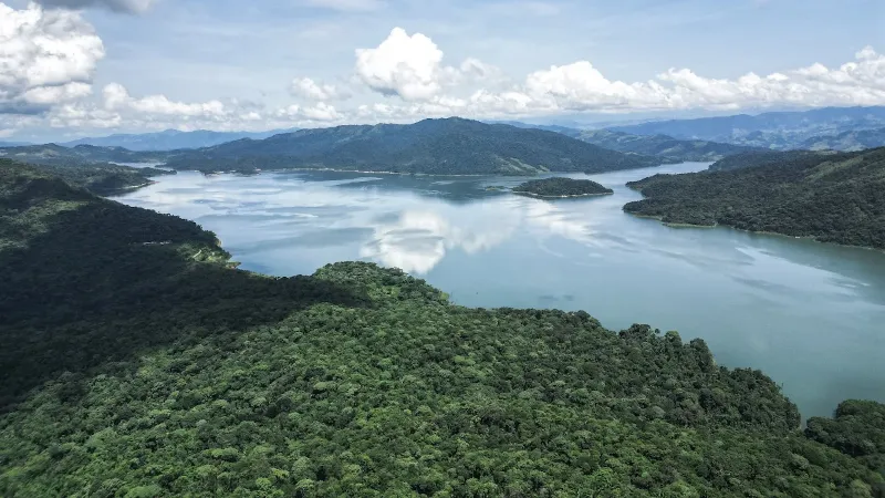 Vista aérea del embalse rodeado de colinas boscosas y reflejos de nubes en el agua bajo cielo despejado