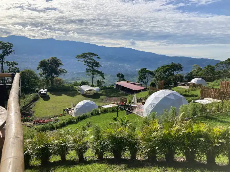 Vista aérea del complejo Santa Cruz Valle Glamping con cuatro domos geodésicos blancos distribuidos en ladera verde, jardines y cordillera al fondo