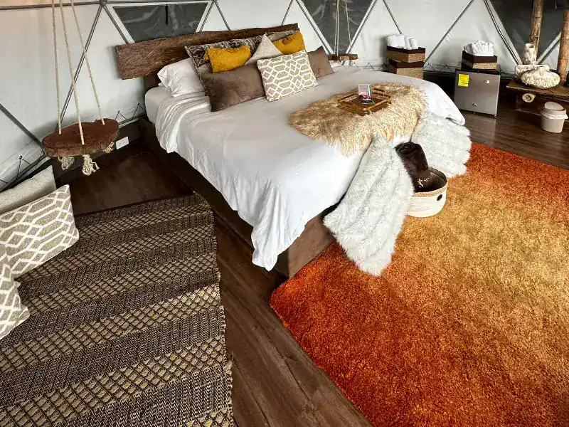 Interior de domo con cama doble de cabecera rústica en madera, cojines dorados, tapete terracota y mesita colgante de cuerda