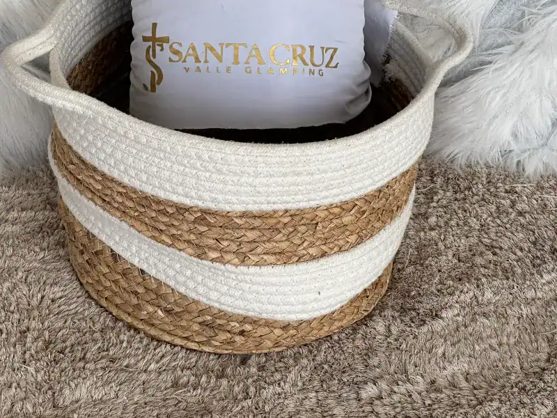 Canasta de fibra natural con almohada blanca bordada con el logo de Santa Cruz Valle Glamping en letras doradas