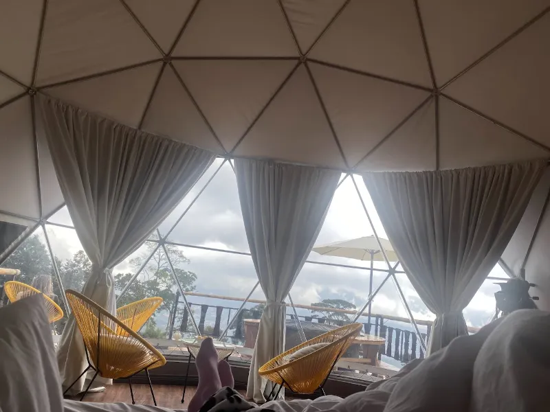 Vista desde la cama del domo hacia el ventanal geodésico con cortinas blancas, sillas naranjas tipo Acapulco y paisaje nublado