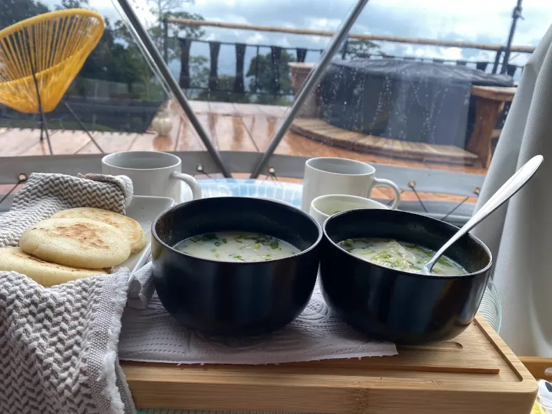 Desayuno en bandeja de madera con dos tazones de caldo, arepas y tazas de café, servido dentro del domo con vista al deck