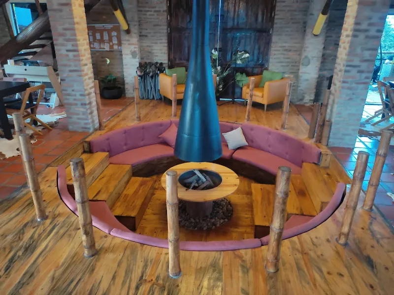 Área social con sofá circular rosa alrededor de chimenea central cónica, pilares de bambú y piso de madera en construcción de ladrillo