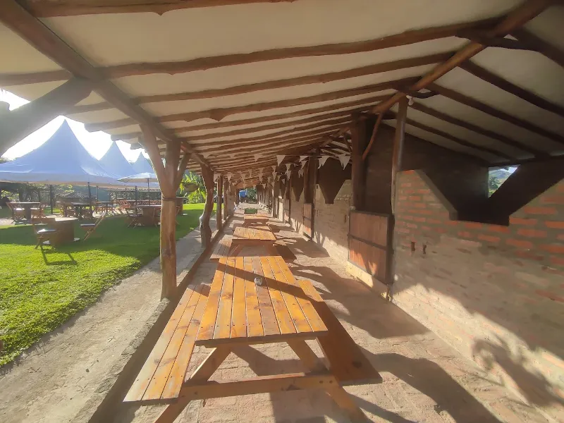 Corredor cubierto con techo de madera y lona, mesas largas de picnic al lado de pared de ladrillo, con jardín verde al fondo