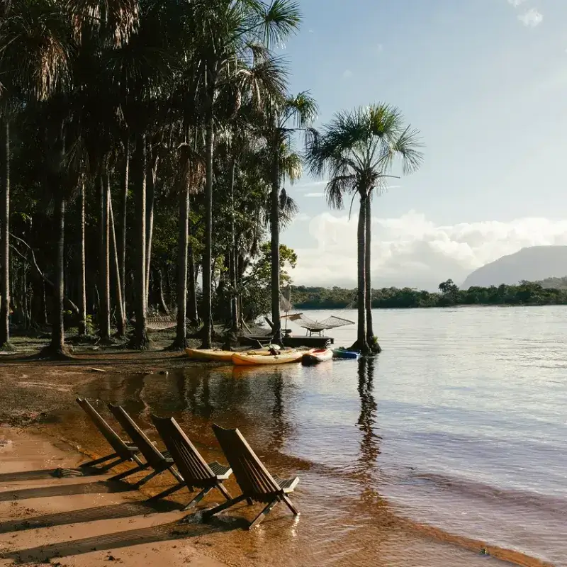Orilla de río con sillas de madera alineadas en la arena frente al agua tranquila, kayaks coloridos y palmeras altas reflejadas en el río, con montañas al fondo en Waku Lodge, Canaima Venezuela.