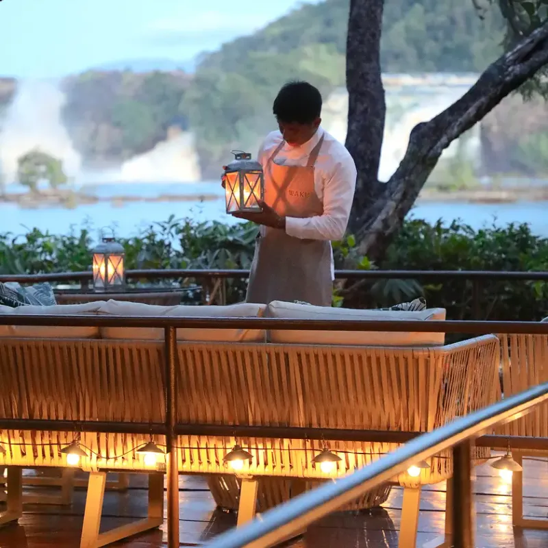 Miembro del personal de Waku Lodge con delantal sosteniendo un farol encendido mientras prepara la terraza exterior al atardecer, con sofás de ratán iluminados por luces cálidas y las cataratas de Canaima visibles al fondo en Waku Lodge, Canaima Venezuela.