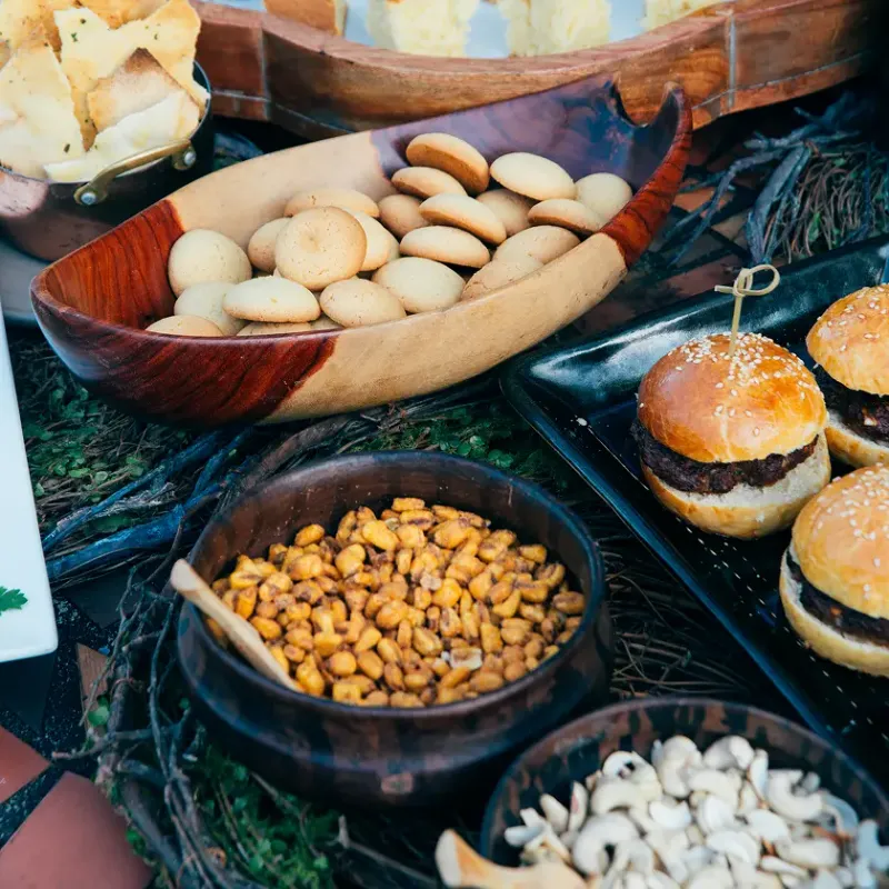 Mesa de aperitivos y bocados gourmet con mini hamburguesas en bandeja negra, galletas en recipiente de madera tallada, maíz tostado y nueces de marañón, presentados sobre una decoración natural con ramas y musgo en Waku Lodge, Canaima Venezuela.