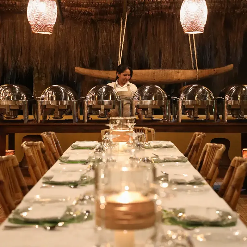Salón comedor elegante con techo de paja artesanal y canoa decorativa suspendida, mesa larga con mantel blanco, vajilla fina, velas y sillas de bambú, con una fila de fuentes de acero inoxidable tipo buffet atendidas por una empleada uniformada bajo lámparas de mimbre rosado en Waku Lodge, Canaima Venezuela.
