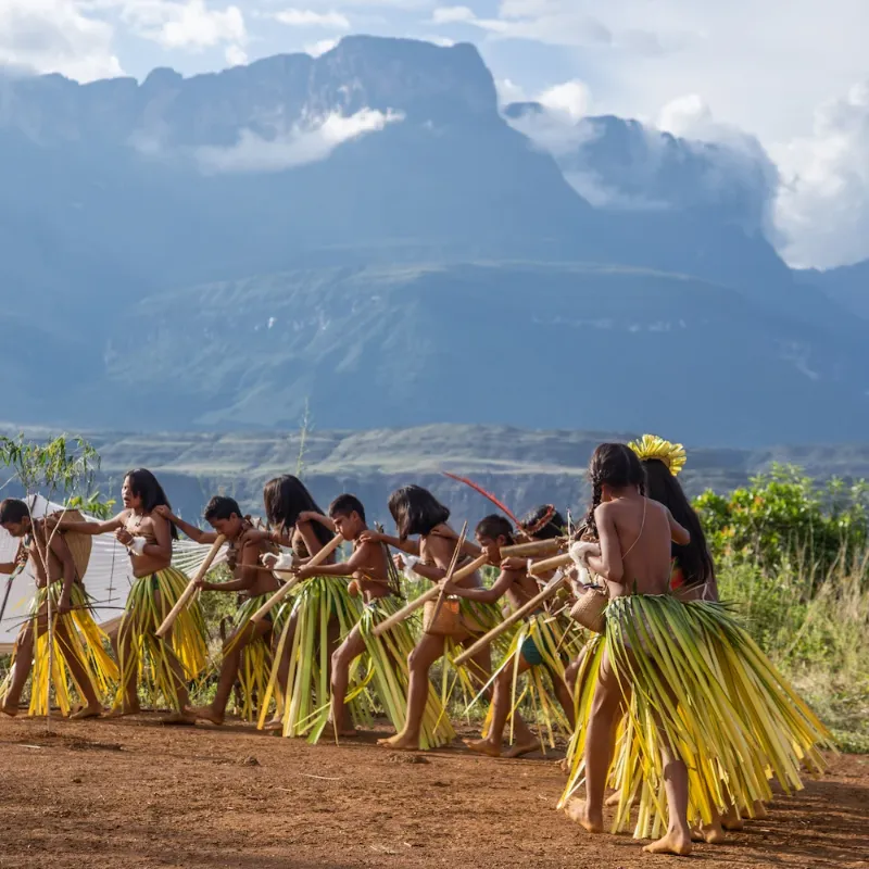 Grupo de niños y jóvenes indígenas pemón con faldas de hojas de palma y accesorios tradicionales realizando una danza ceremonial en tierra, con el imponente tepuy Roraima al fondo entre nubes en Waku Lodge, Canaima Venezuela.
